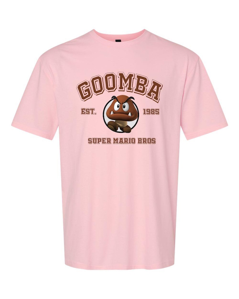 A pink t-shirt with a graphic of Goomba, a character from the Super Mario Bros. video game franchise, and text that reads "GOOMBA EST. 1985 SUPER MARIO BROS".