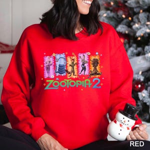 A woman wearing a red sweater with a colorful graphic design featuring characters from the movie "Zootopia 2". She is holding a small snowman toy.