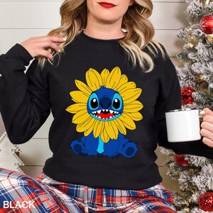 A woman wearing a black sweatshirt with a cartoon character of a blue cat with a yellow flower on it, holding a white mug.