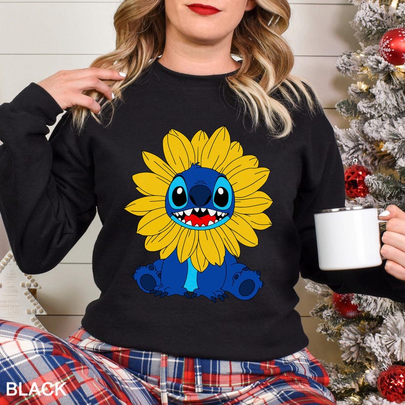 A woman wearing a black sweatshirt with a cartoon character of a blue cat with a yellow flower on it, holding a white mug.