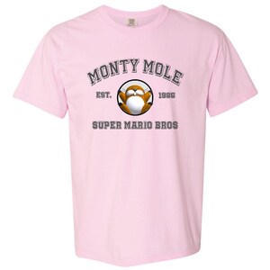 A pink t-shirt with a graphic design featuring a cartoon character and text that reads "MONTY MOLE EST. 1985 SUPER MARIO BROS".