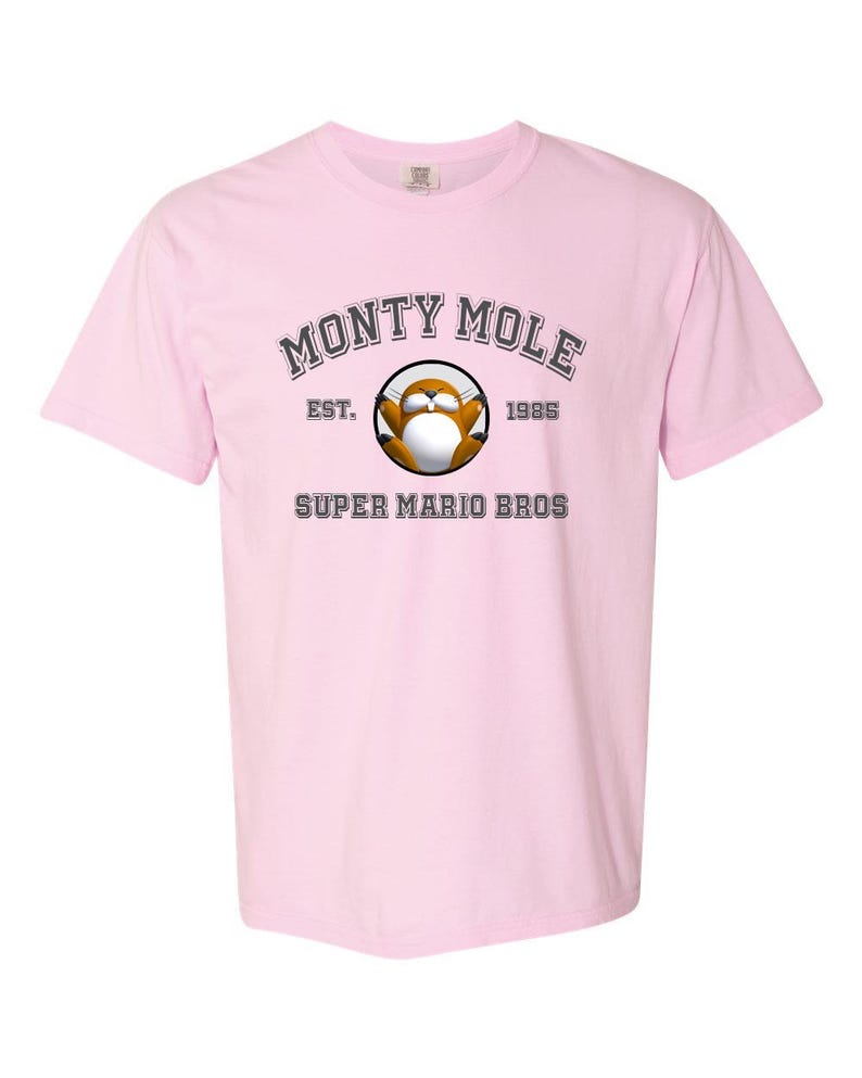 A pink t-shirt with a graphic design featuring a cartoon character and text that reads "MONTY MOLE EST. 1985 SUPER MARIO BROS".