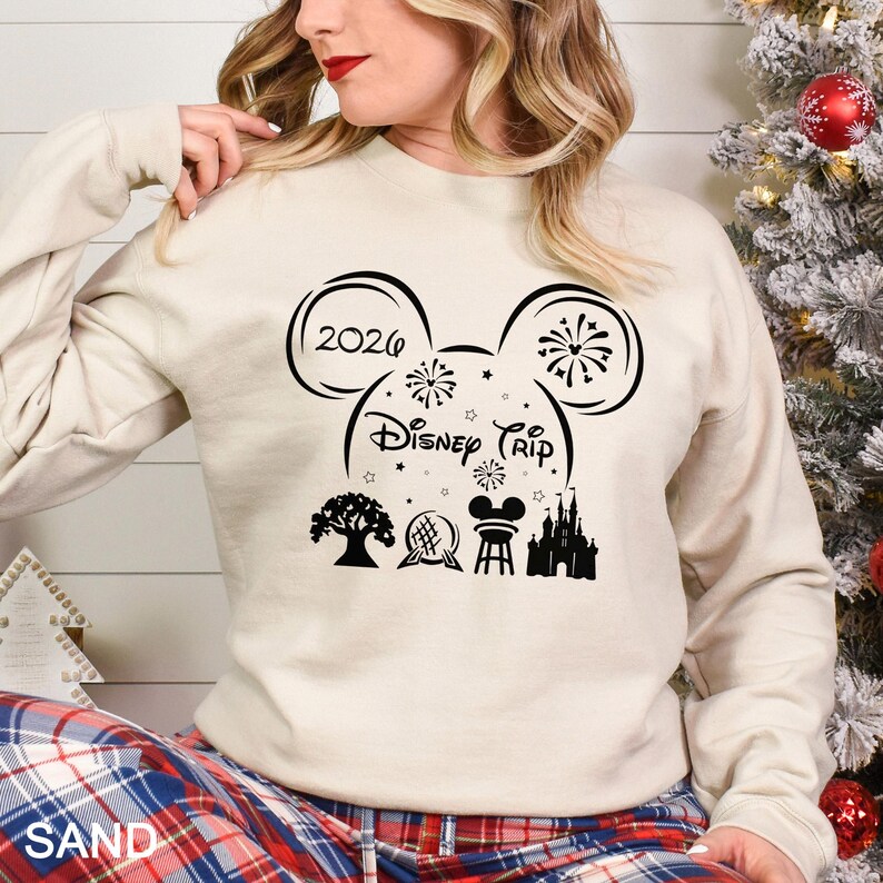 A woman wearing a white sweatshirt with a graphic design featuring Disney-themed elements such as Mickey Mouse ears, fireworks, and a castle.