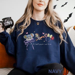 A woman wearing a navy blue sweatshirt with a graphic design of cartoon characters and text, holding a black mug with a Halloween-themed design.