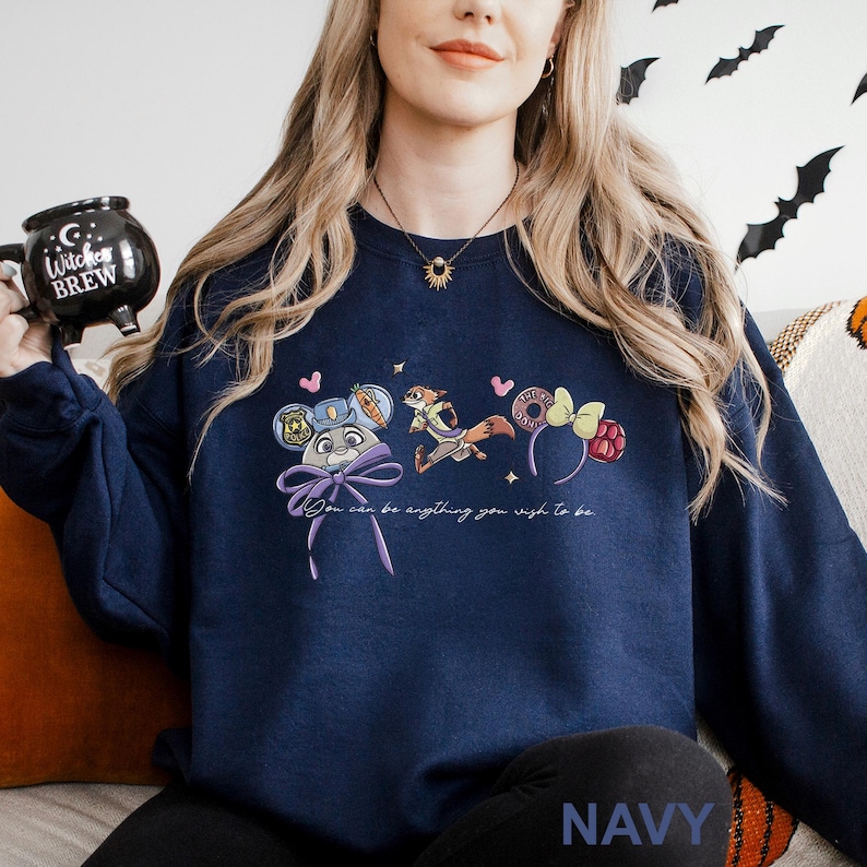 A woman wearing a navy blue sweatshirt with a graphic design of cartoon characters and text, holding a black mug with a Halloween-themed design.