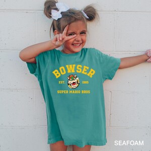 A young girl wearing a green t-shirt with a Mario Bros. logo and text, smiling and making a peace sign gesture.