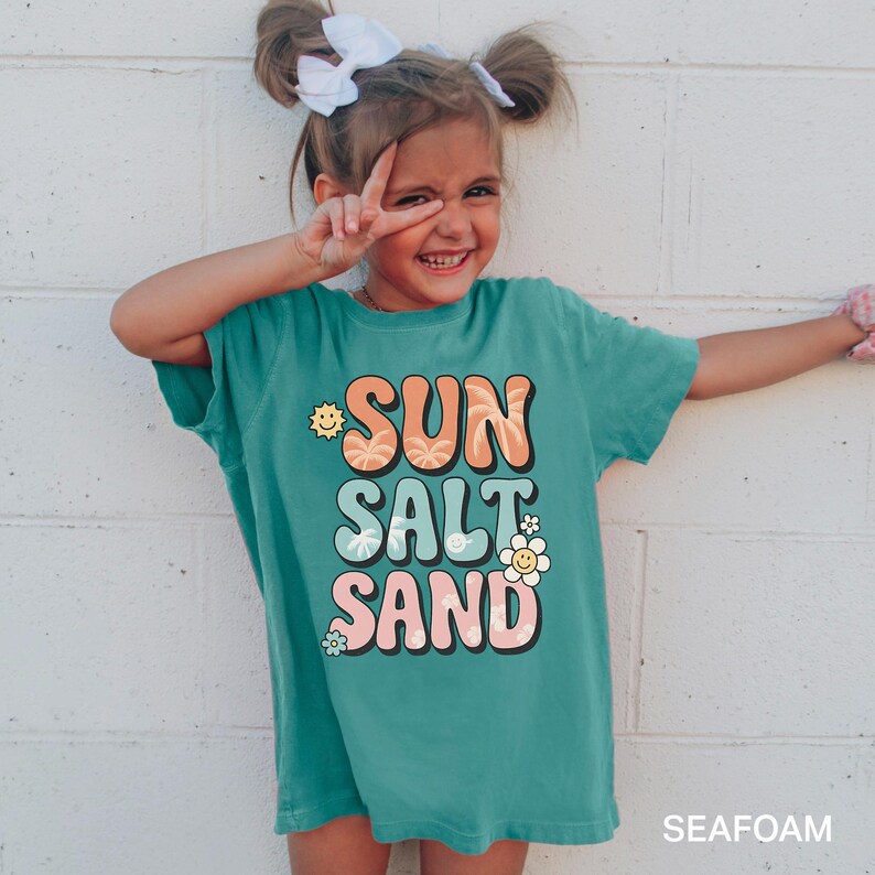 A young girl with pigtails is smiling and making a peace sign with her fingers, wearing a green t-shirt with the text "SUN SALT SAND" printed on it.