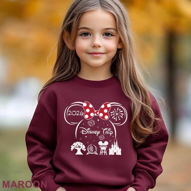 A young girl with long blonde hair wearing a maroon sweater with a Disney-themed design and text.