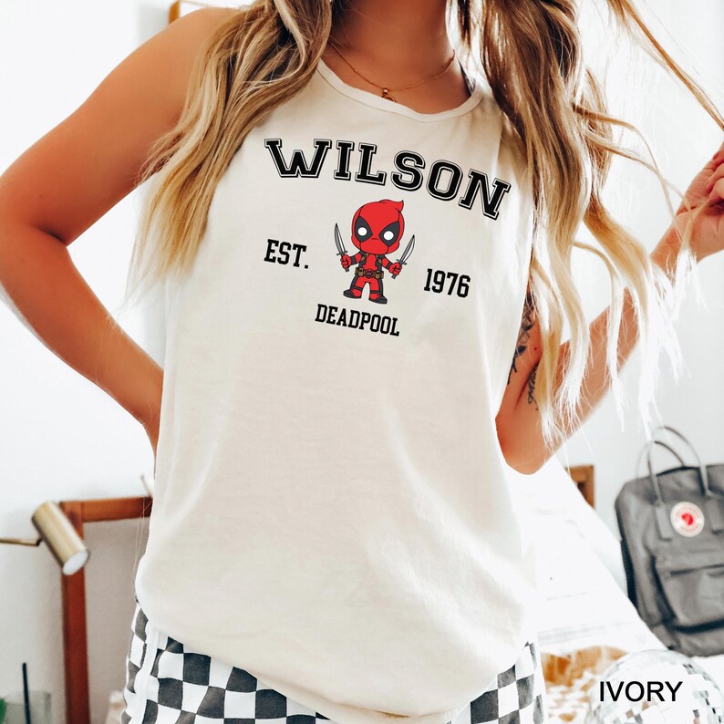 A woman wearing a white tank top with a graphic of a character from the Marvel Cinematic Universe, specifically Deadpool.