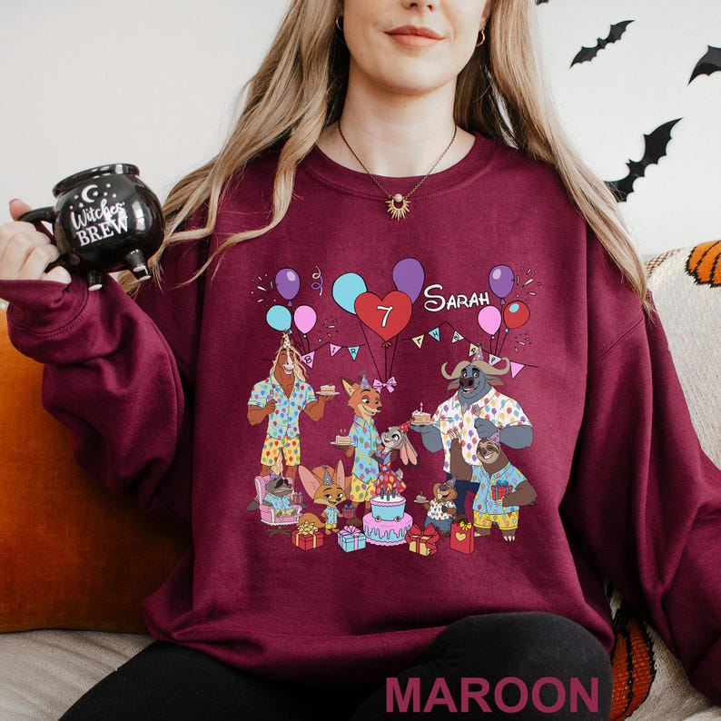A woman wearing a maroon sweatshirt with a graphic design of cartoon animals and a birthday cake, holding a black mug with the text "COOK BREW" on it.