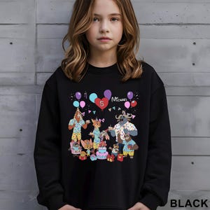A young girl wearing a black sweatshirt with a colorful illustration of cartoon animals and a birthday party scene.