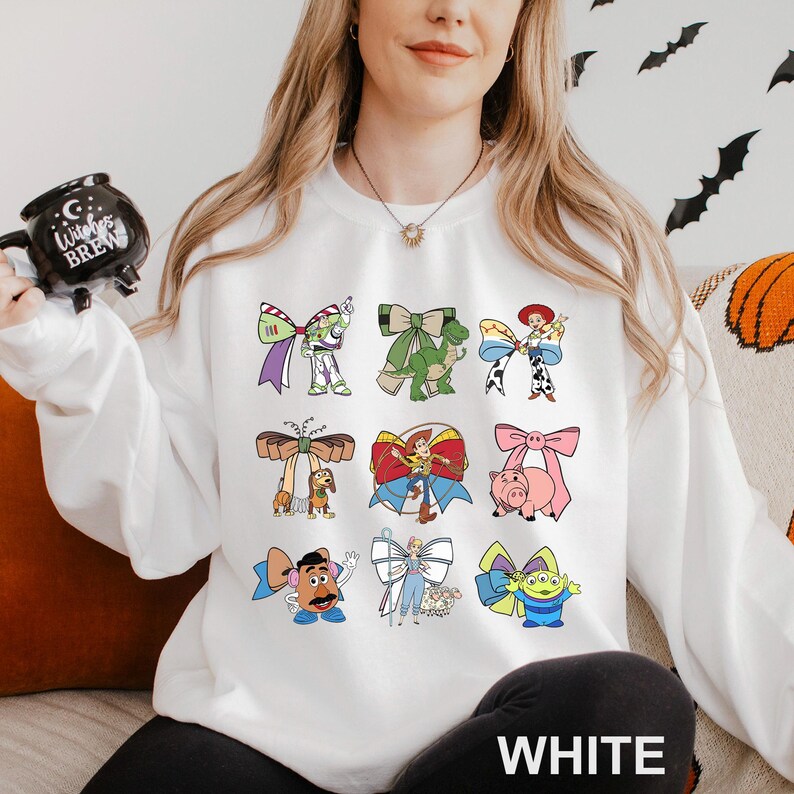 A woman wearing a white sweatshirt with a collage of cartoon characters from Toy Story printed on it, holding a mug with a black design.