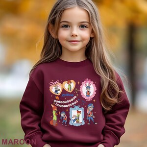 A young girl with long blonde hair wearing a maroon sweater with various cartoon characters and designs.