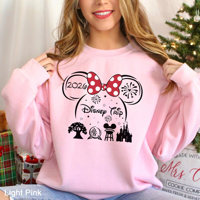 A woman wearing a pink sweatshirt with a Disney-themed design, including a red bow, fireworks, and various Disney landmarks.