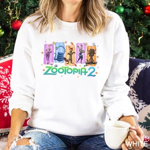A woman wearing a white sweatshirt with a colorful graphic of five cartoon animal characters from the movie "Zootopia 2" on it, sitting on a couch and holding a white mug.