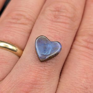 May include: A small heart-shaped gemstone with a smooth, polished surface displaying iridescent blue and violet hues. The stone features a dark, natural-looking base and is positioned on a finger next to a plain, polished gold band ring.