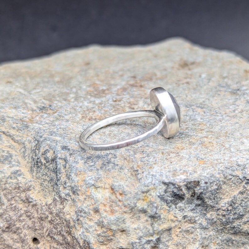 Op de afbeelding: Een zilveren ring met een grijze edelsteen. De ring heeft een gehamerde textuur en ligt op een grijze steen.