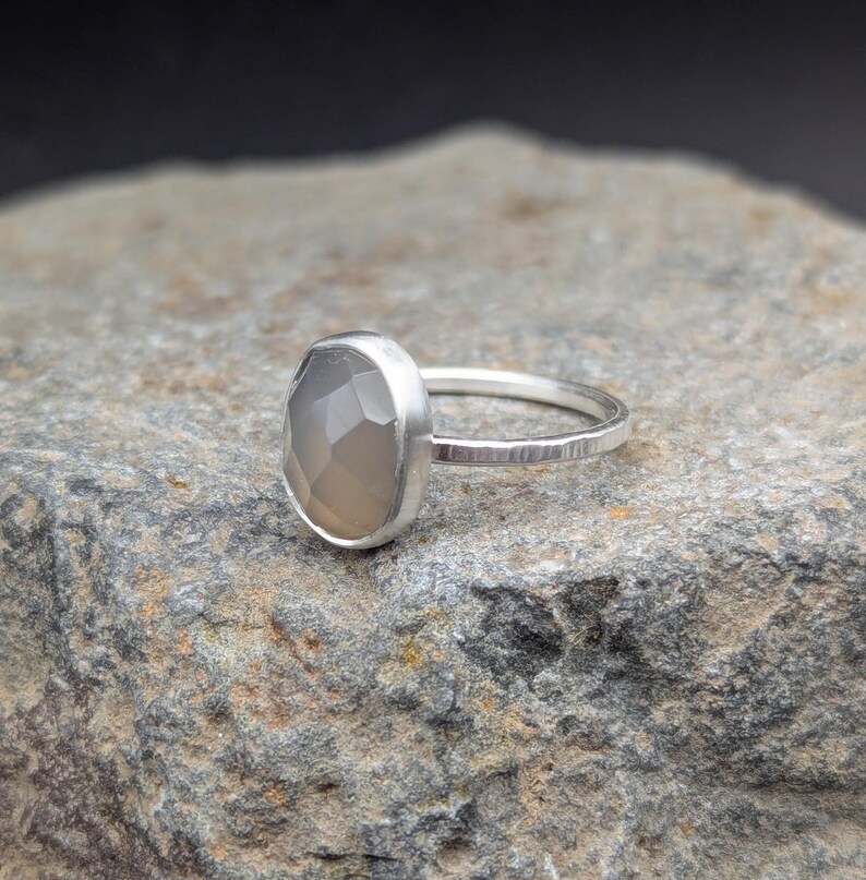 Op de afbeelding: Een zilveren ring met een grijze edelsteen in een bezel-setting. De ring ligt op een grijze rots.