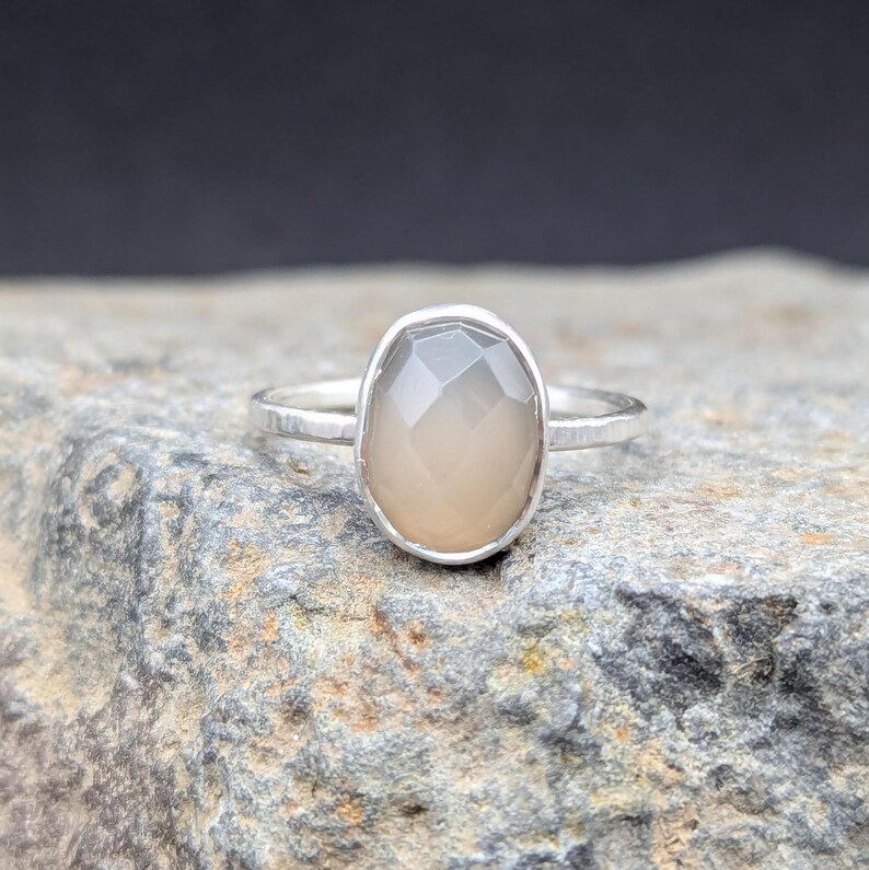 Op de afbeelding: Een zilveren ring met een lichtgrijze ovale edelsteen. De ring heeft een gehamerde textuur.
