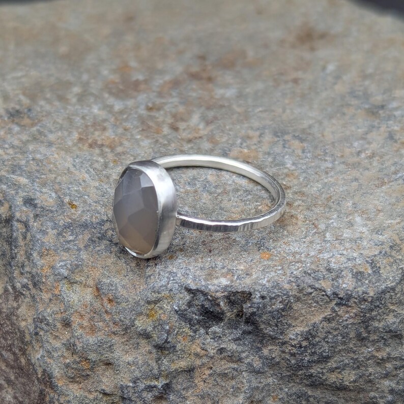 Op de afbeelding: Een zilveren ring met een grijze ovale edelsteen. De ring heeft een getextureerde band.