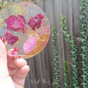 May include: A round, clear resin coaster with dried pink and red flower petals and gold glitter embedded inside.