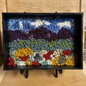 May include: A framed fiber art piece depicting a landscape scene. The artwork features a textured, colorful design with mountains, sky, and foliage. The frame is black and has a stand.