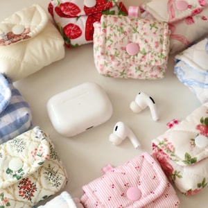 May include: A collection of small, quilted fabric pouches with snap closures, featuring various floral, gingham, and strawberry patterns, arranged around a white wireless earbud charging case and a pair of white wireless earbuds.