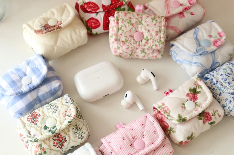 Pu&ograve; includere: Una collezione di piccole pochette in tessuto trapuntato con chiusure a scatto, caratterizzate da vari motivi floreali, a quadretti e a fragole, disposte attorno a una custodia di ricarica per auricolari wireless e un paio di auricolari bianchi.