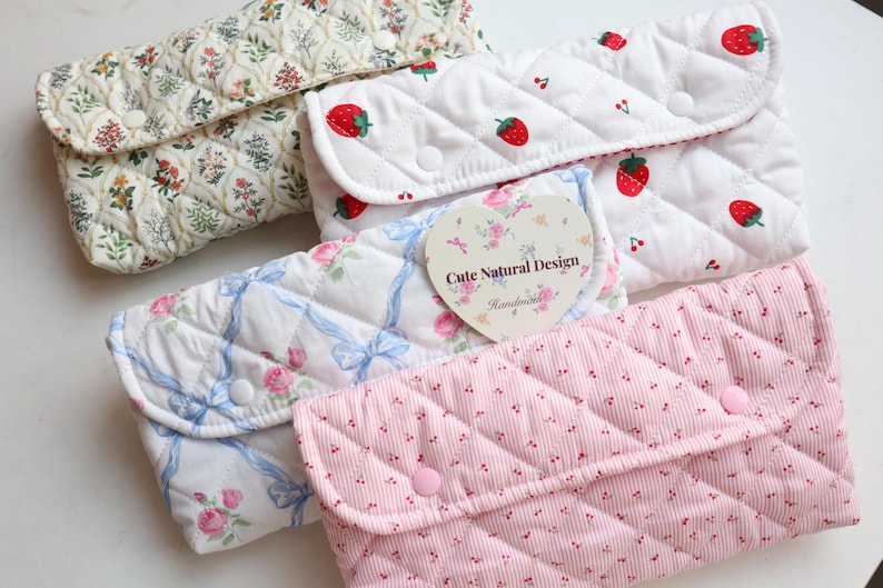 May include: A collection of quilted fabric pouches in various patterns. Designs include floral, strawberries, roses with blue ribbons, and a pink striped pattern. Each pouch has a button closure.