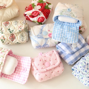 May include: A collection of small, quilted fabric cases in various floral and patterned designs. Some cases are open, revealing white earbud cases. The cases feature button closures and a variety of colors, including pink, blue, and white.