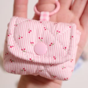 Pu&ograve; includere: Una piccola custodia trapuntata a righe rosa chiaro e bianche con un motivo a ciliegie ripetuto. La custodia presenta una patta chiusa da un bottone a pressione rosa e un anello in plastica rosa fissato tramite una linguetta a quadretti.