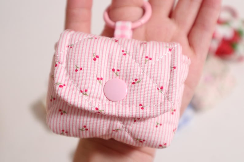 Pu&ograve; includere: Una piccola custodia trapuntata a righe rosa chiaro e bianche con un motivo a ciliegie ripetuto. La custodia presenta una patta chiusa da un bottone a pressione rosa e un anello in plastica rosa fissato tramite una linguetta a quadretti.