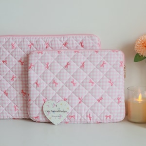 May include: Two pink quilted laptop sleeves with a gingham pattern and embroidered bows. The sleeves have a pink zipper and are in different sizes. A heart-shaped tag reads "Cute Natural Design Handmade."