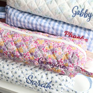 May include: A stack of quilted pencil cases in various floral and patterned fabrics. The cases are embroidered with names like "Gabby", "Phoebe", "Georgine", and "Sarah". The cases have a zipper closure and a small handle.