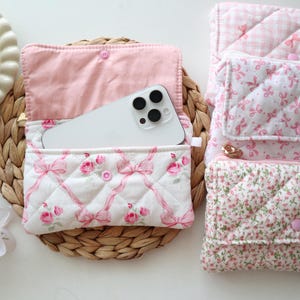 May include: A collection of four rectangular quilted fabric pouches featuring various pink and white patterns including floral prints, bows, and gingham checks. Each pouch has a flap closure with a pink snap button and a soft interior lining.