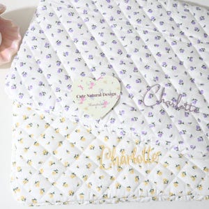 May include: A quilted laptop sleeve featuring a white fabric with a small purple floral pattern and a separate white fabric with a small yellow floral pattern. The name Charlotte is embroidered in purple and gold thread. A heart-shaped tag reads Cute Natural Design.