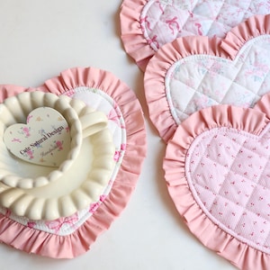 May include: Heart-shaped quilted placemats with ruffled edges in pink. One placemat has a cream-colored bowl and a heart-shaped tag that reads "Cute Natural Design". The placemats are decorated with floral patterns.