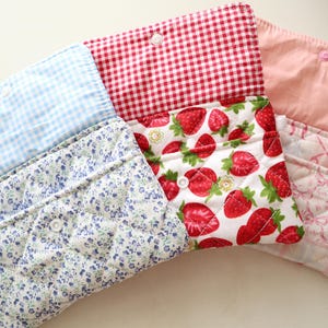 May include: Three quilted fabric pouches with snap closures. The first features a blue gingham flap and blue floral print body. The second has a red gingham flap and strawberry print body. The third has a solid pink flap and pink plaid body.