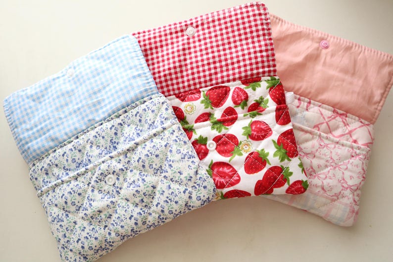 May include: Three quilted fabric pouches with snap closures. The first features a blue gingham flap and blue floral print body. The second has a red gingham flap and strawberry print body. The third has a solid pink flap and pink plaid body.