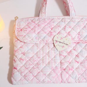 May include: A quilted pink and white tote bag with a floral and bow pattern. The bag has a flap closure and a heart-shaped tag that reads "Cute Natural Design". The bag has a top handle and a zipper closure.