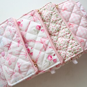 May include: A quilted fabric organizer featuring multiple sections with pink and white patterns, including floral prints, ribbons, and gingham checks. The organizer has pink zippers, snap closures, and a soft, padded texture throughout the design.