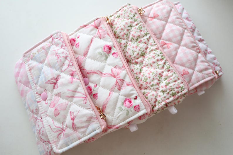 May include: A quilted fabric organizer featuring multiple sections with pink and white patterns, including floral prints, ribbons, and gingham checks. The organizer has pink zippers, snap closures, and a soft, padded texture throughout the design.