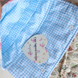 May include: Close-up of a handmade fabric item with a blue and white gingham interior. The item has a floral print exterior and a heart-shaped tag that reads "Cute Natural Design Handmade."