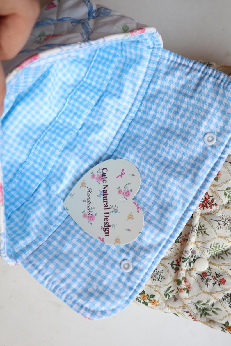 May include: Close-up of a handmade fabric item with a blue and white gingham interior. The item has a floral print exterior and a heart-shaped tag that reads "Cute Natural Design Handmade."