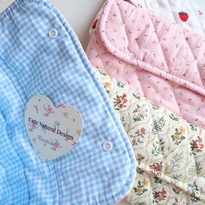 May include: Close-up of three fabric pouches in pastel colors. One is light blue gingham, another is pink quilted with small dots, and the third is cream with a floral pattern. Each has a snap closure. A heart-shaped tag reads "Cute Natural Design".