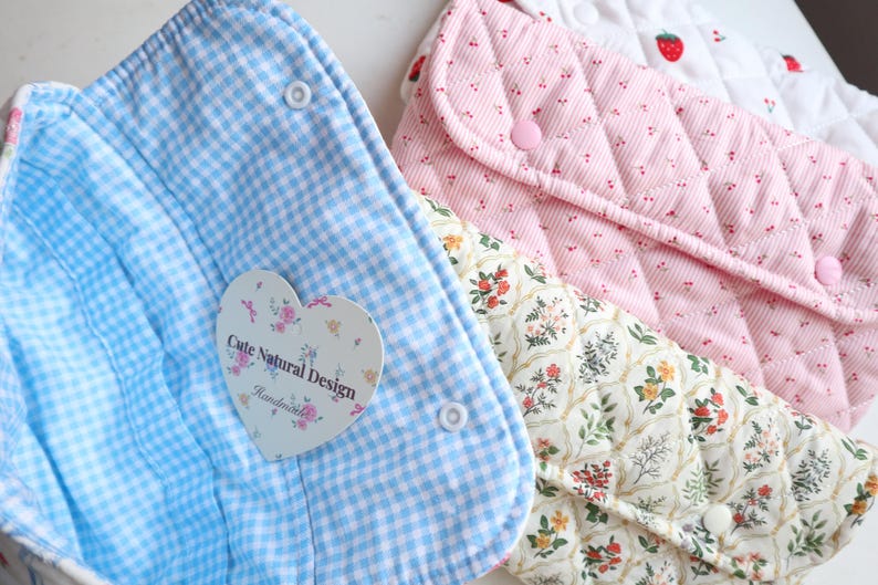 May include: Close-up of three fabric pouches in pastel colors. One is light blue gingham, another is pink quilted with small dots, and the third is cream with a floral pattern. Each has a snap closure. A heart-shaped tag reads "Cute Natural Design".