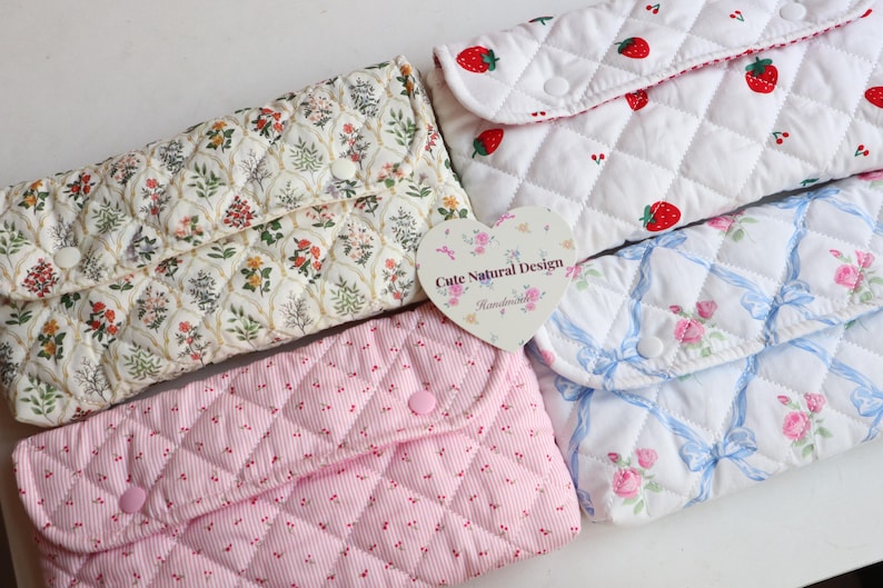 May include: A collection of quilted fabric pouches in various patterns. Designs include floral, strawberries, and roses with bows. The pouches have a button closure. A heart-shaped tag reads "Cute Natural Design".