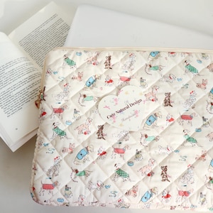 May include: A quilted laptop sleeve with a repeating pattern of dogs in various outfits. The sleeve is cream-colored with a diamond pattern. A heart-shaped tag reads "Cute Natural Design". An open book is visible.