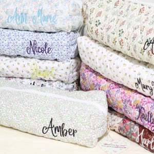 May include: A stack of personalized floral print pillows in various colors, including blue, white, and pink. Each pillow is embroidered with a name in a cursive font. The pillows are quilted and have a soft, inviting appearance.