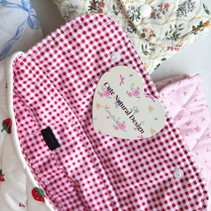 May include: Close-up of several quilted fabric pouches with different patterns. One features a red and white gingham lining, another has a floral design, and a third has a strawberry pattern. A heart-shaped tag reads "Cute Natural Design Handmade."